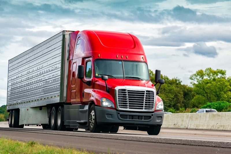 A large red semi-truck drives down a highway during a cloudy day.