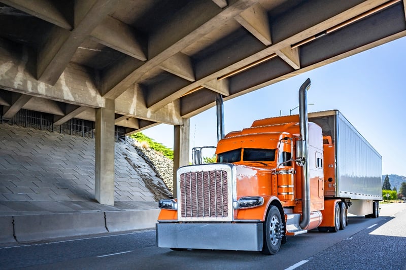 An orange semi-truck drives under an overpass on a highway. 
