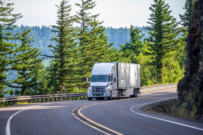 A semi-truck drives down a scenic mountain highway surrounded by trees.