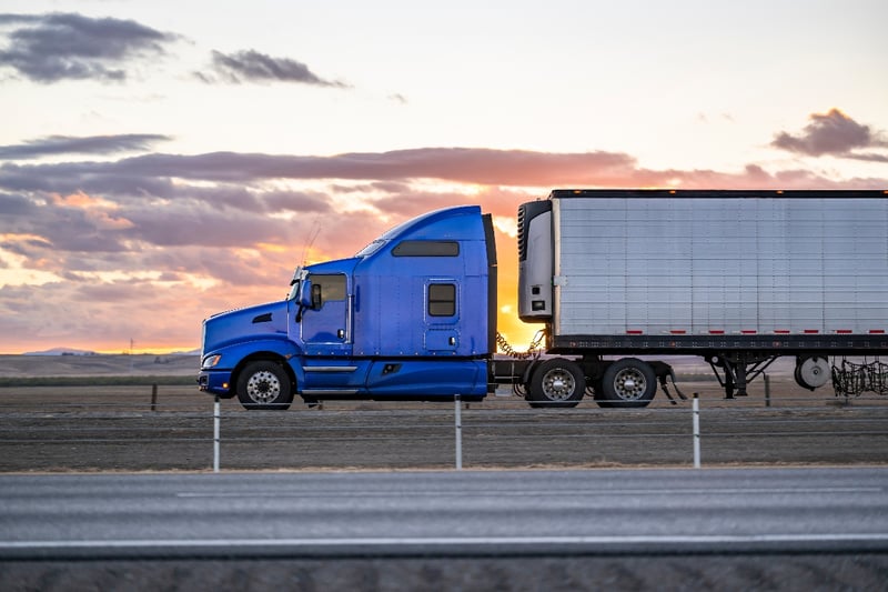 A blue semi-truck drives down a highway at dusk.